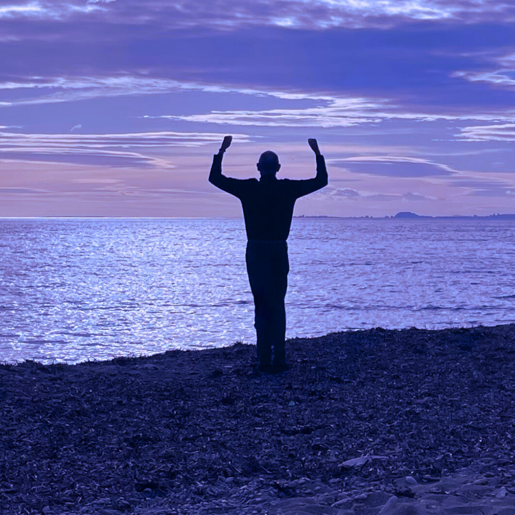 Een man die voor op het strand staat naar de zee te kijken, je ziet hem in silhouet, met zijn armen gebald naar boven, met de bedoeling van sta in je kracht uit te beelden. 